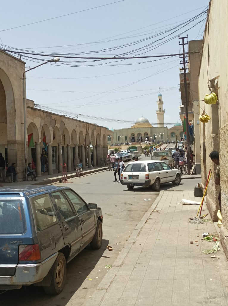 Mala tržnica s lijeva, okružena dućanima i u pozadini se vidi velika džamija Khulafa al-Rashidun.